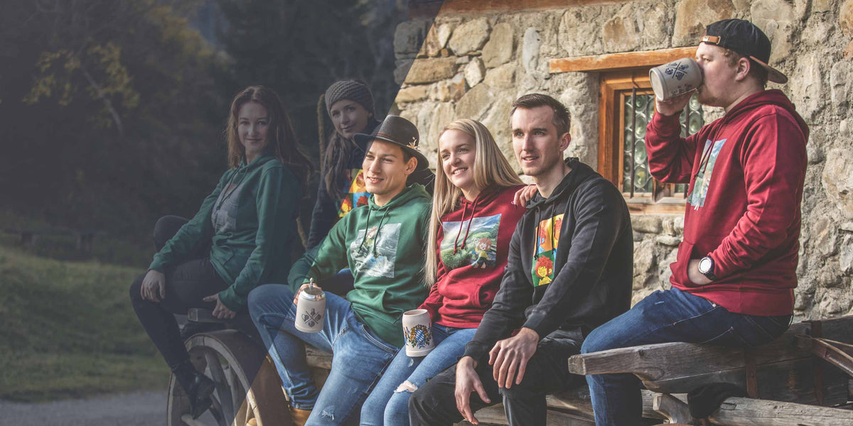 Gruppe von Freunden in Unisex Hoodies mit bayerischen Motiven sitzen auf einer Holzbank vor einem Steinhaus.