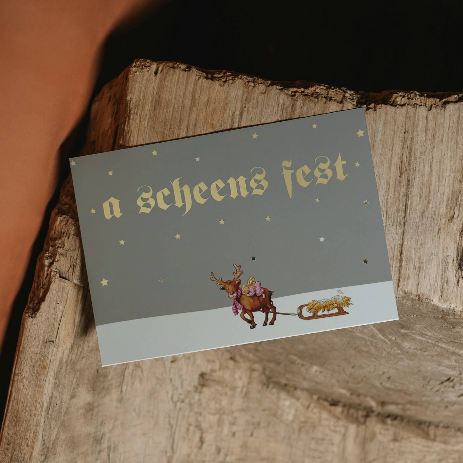 Glückwunschkarte a scheens Fest - Christkind mit Rentier und Goldbuchstaben auf Holzuntergrund.