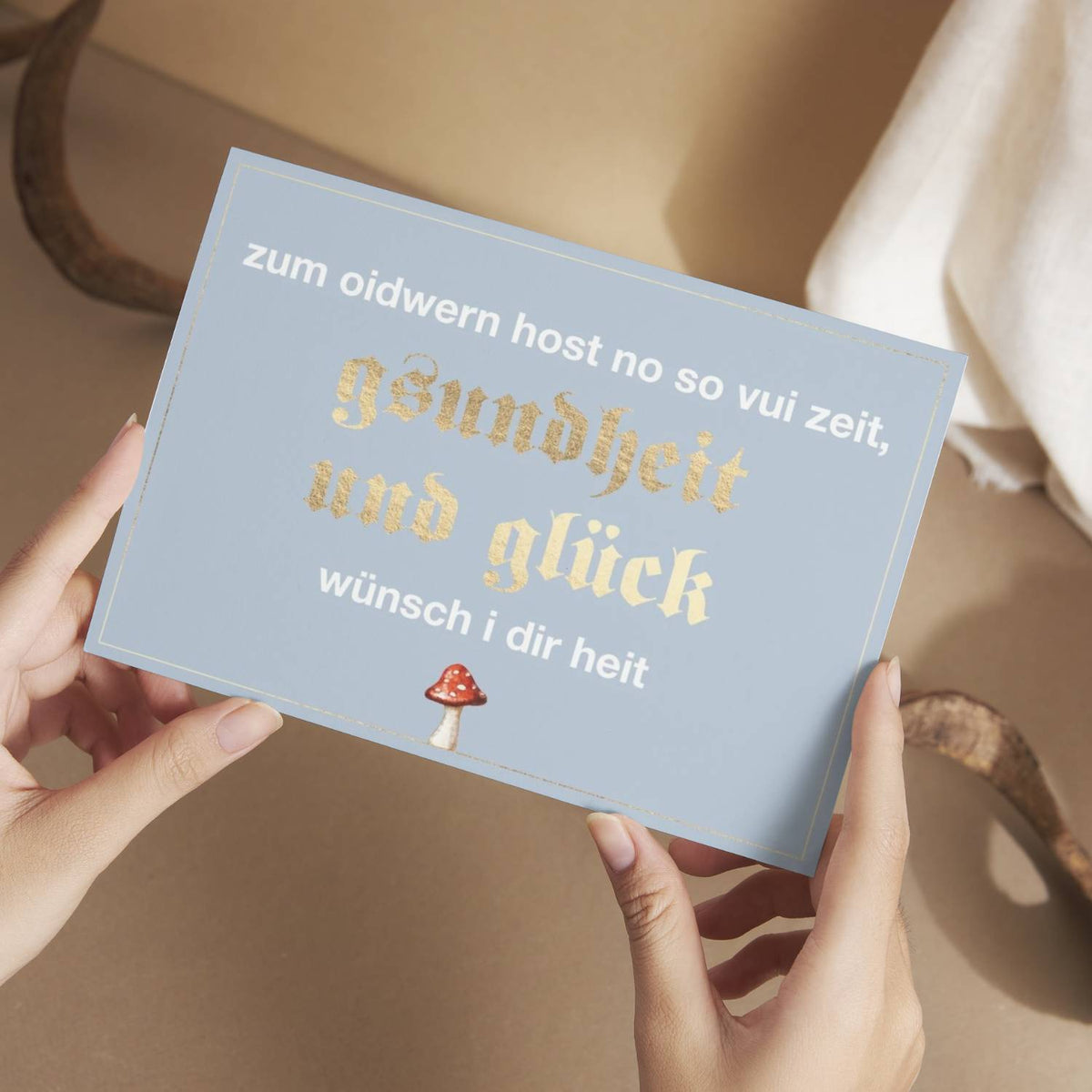 Hände halten die hellblaue Glückwunschkarte "Zum Oidwern...", verziert mit goldener Schrift und einem Fliegenpilz-Motiv.