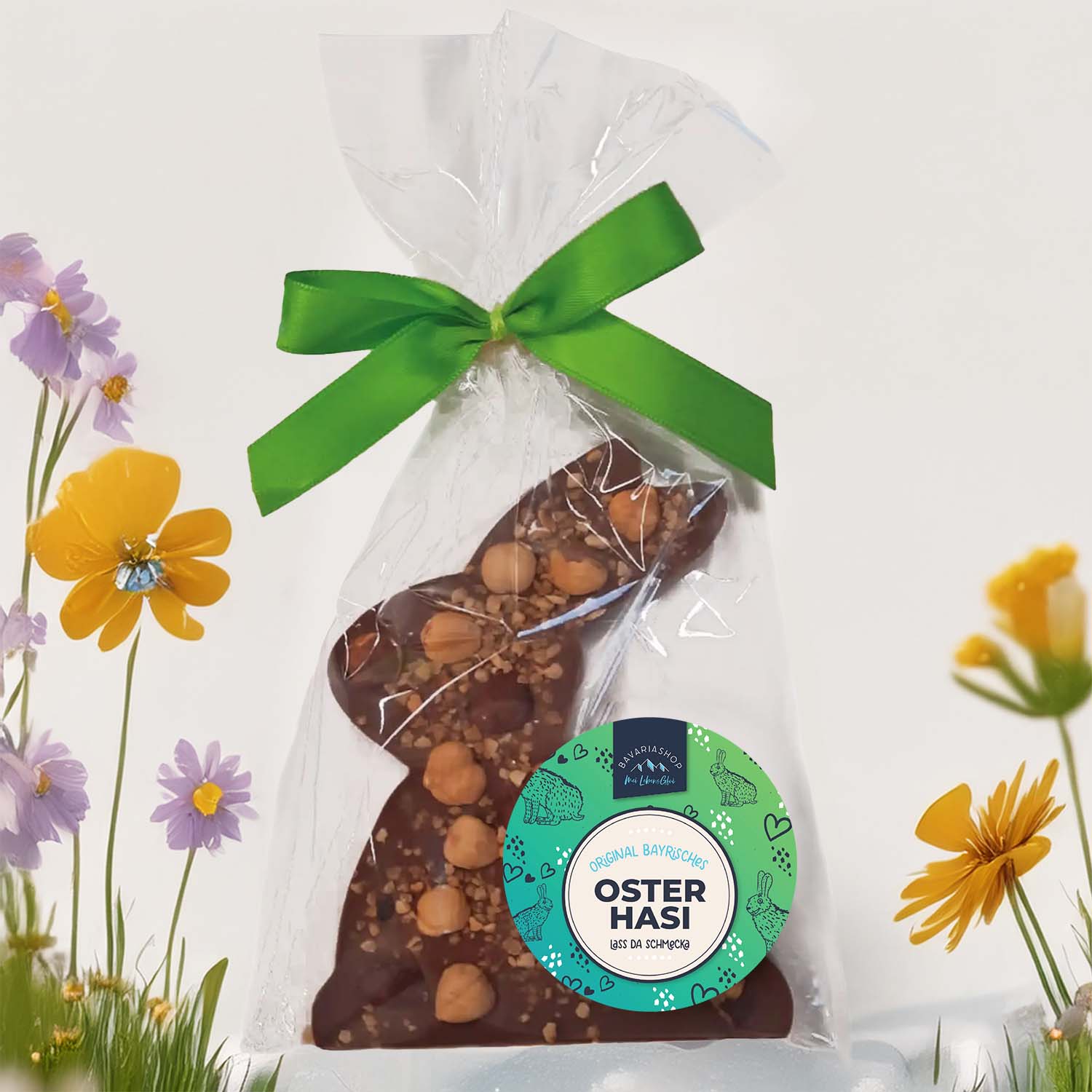 Oster-Schokoladen Nuss-Haserl in Klarsichtverpackung mit grüner Schleife, umgeben von bunten Frühlingsblumen.