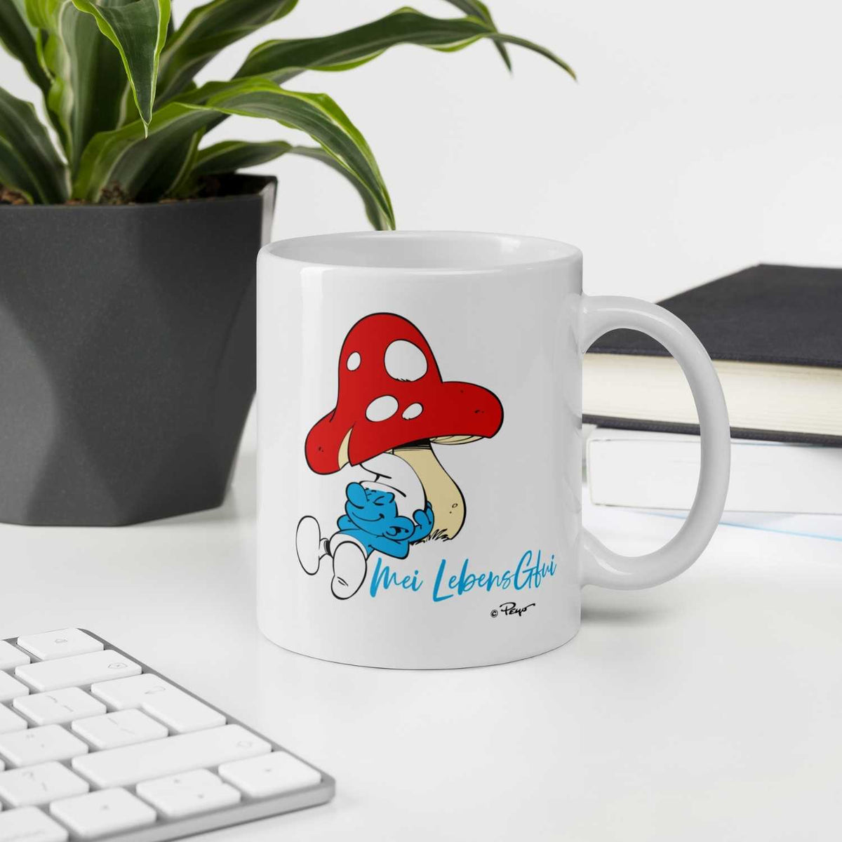 Die Schlümpfe Tasse Do legst di nieda mit lustigem Schlumpf unter rotem Pilz und blauem Schriftzug auf weißem Schreibtisch.