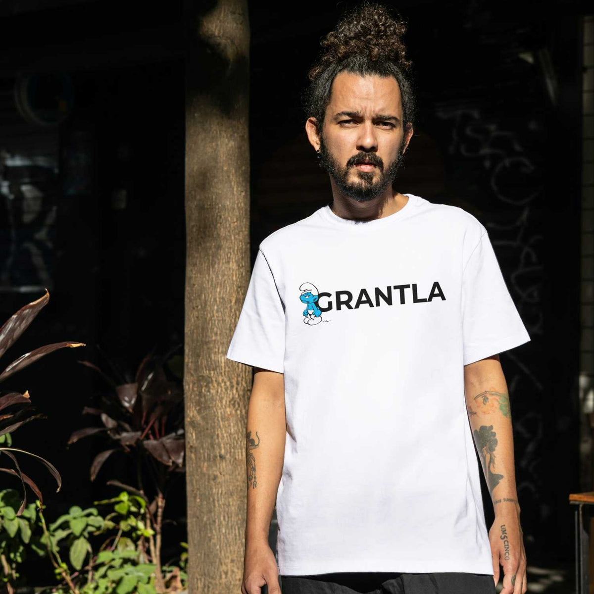 Junger Mann mit Bart und weißem Die Schlümpfe Herren T-Shirt "Grantla" vor dunklem Hintergrund und Baumstamm.