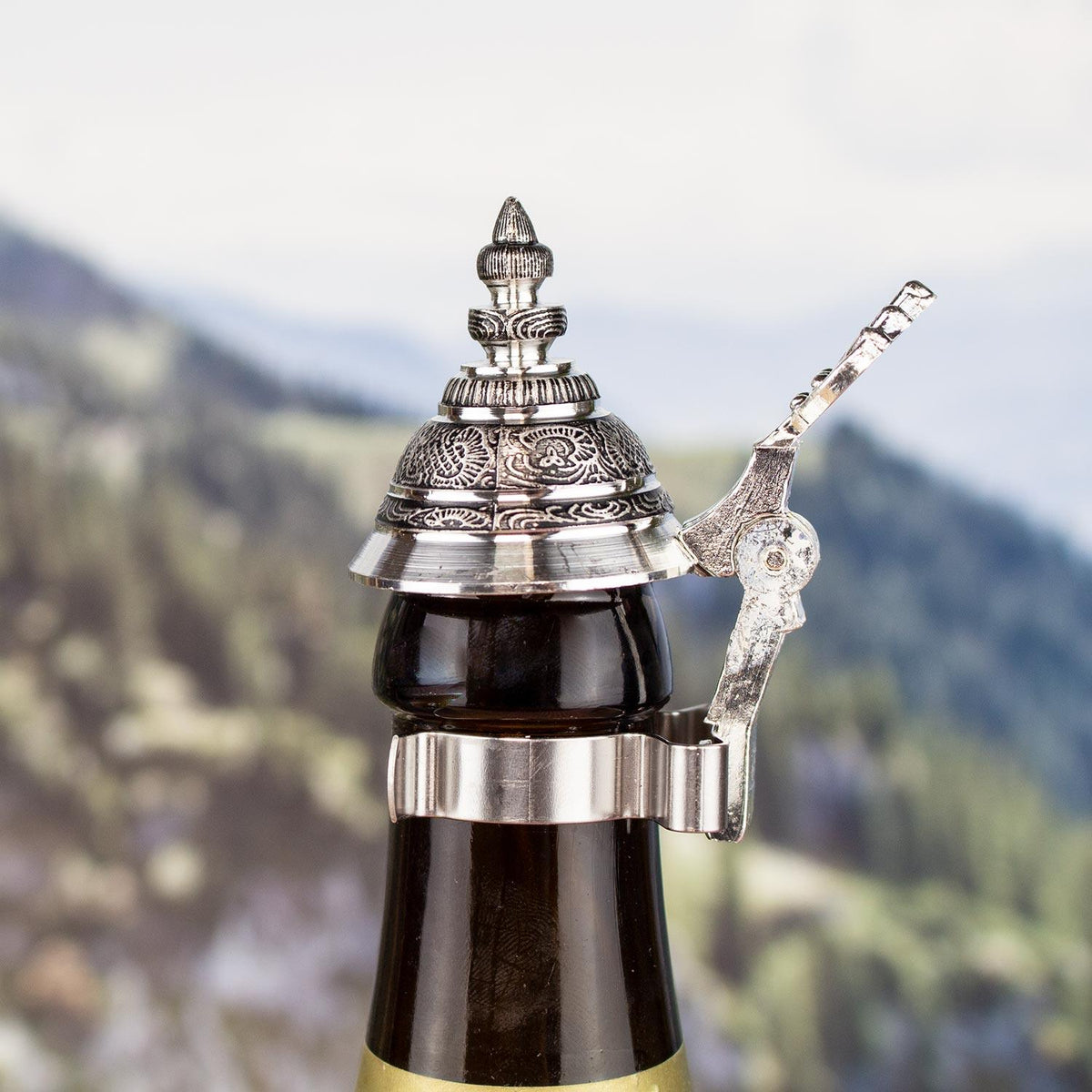 Detailansicht der Geschenkbox "Bierdimpfe" mit verziertem Zinndeckel auf einer Bierflasche vor Alpenkulisse.