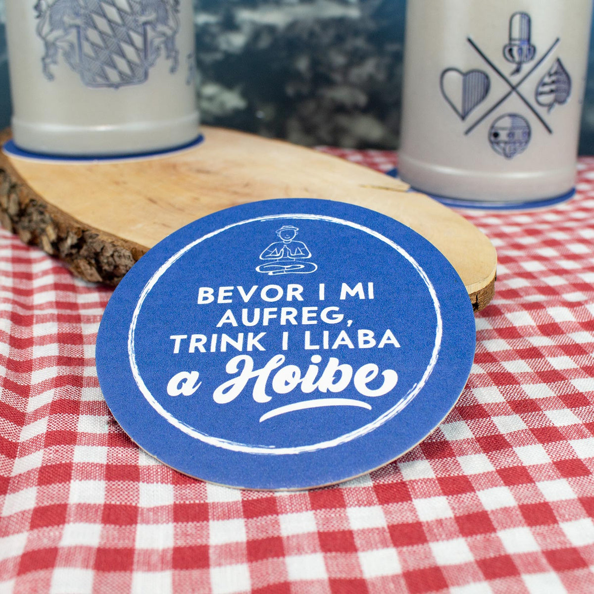 Bierfuizl "Bevor i mi aufreg..." in blau mit weißer Aufschrift auf rot-weiß karierter Tischdecke und Holzbrett.