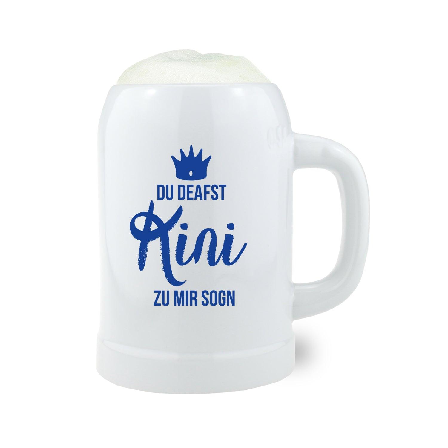 Weißer Bierkrug Du deafst Kini zu mir sogn mit blauem Aufdruck und Krone, gefüllt mit Schaum.