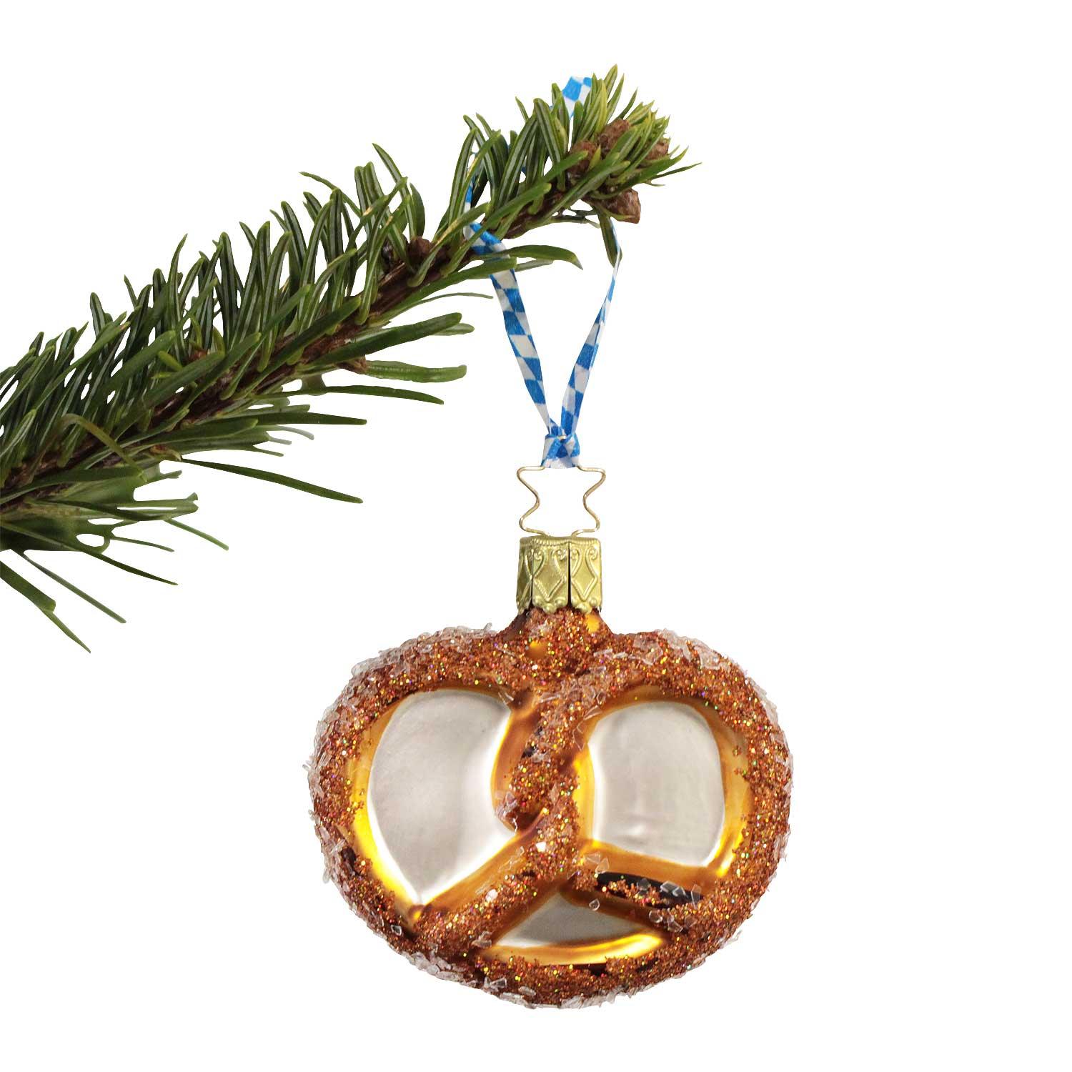 Handgefertigter Weihnachtsbaumschmuck in Form einer glitzernden Brezel hängt an einem Fichtenzweig mit Bayernband.
