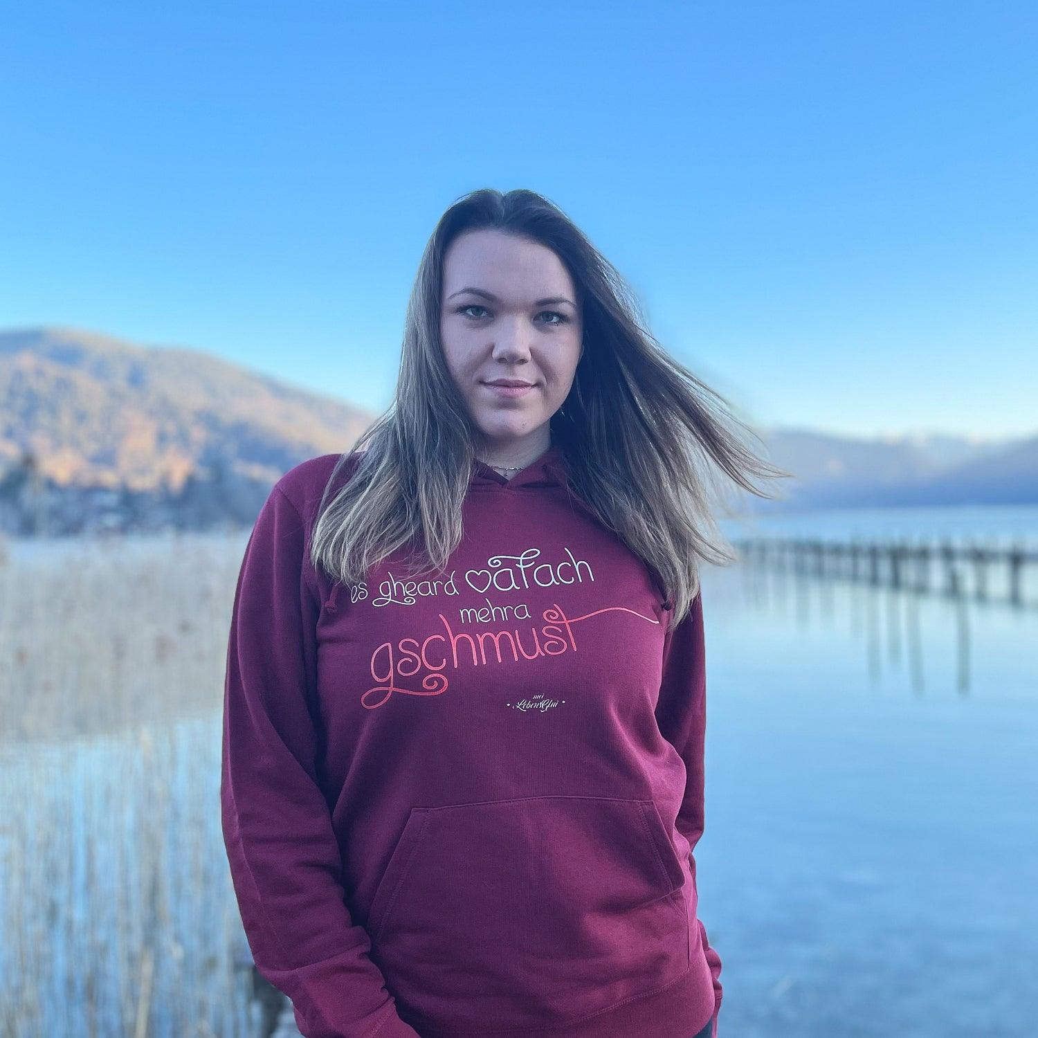 Junge Frau trägt dunkelroten Damen Hoodie Gschmust am Seeufer, im Hintergrund Berge und blauer Himmel.