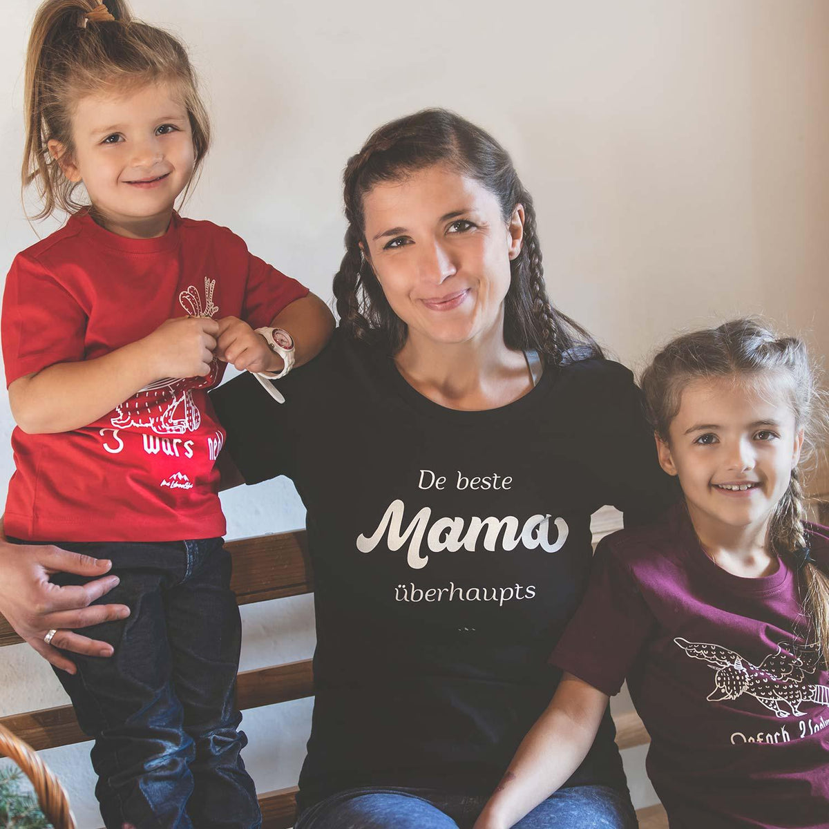 Lächelnde Mutter im schwarzen Damen T-Shirt "De beste Mama übahaupts" mit zwei Kindern auf Holzbank.