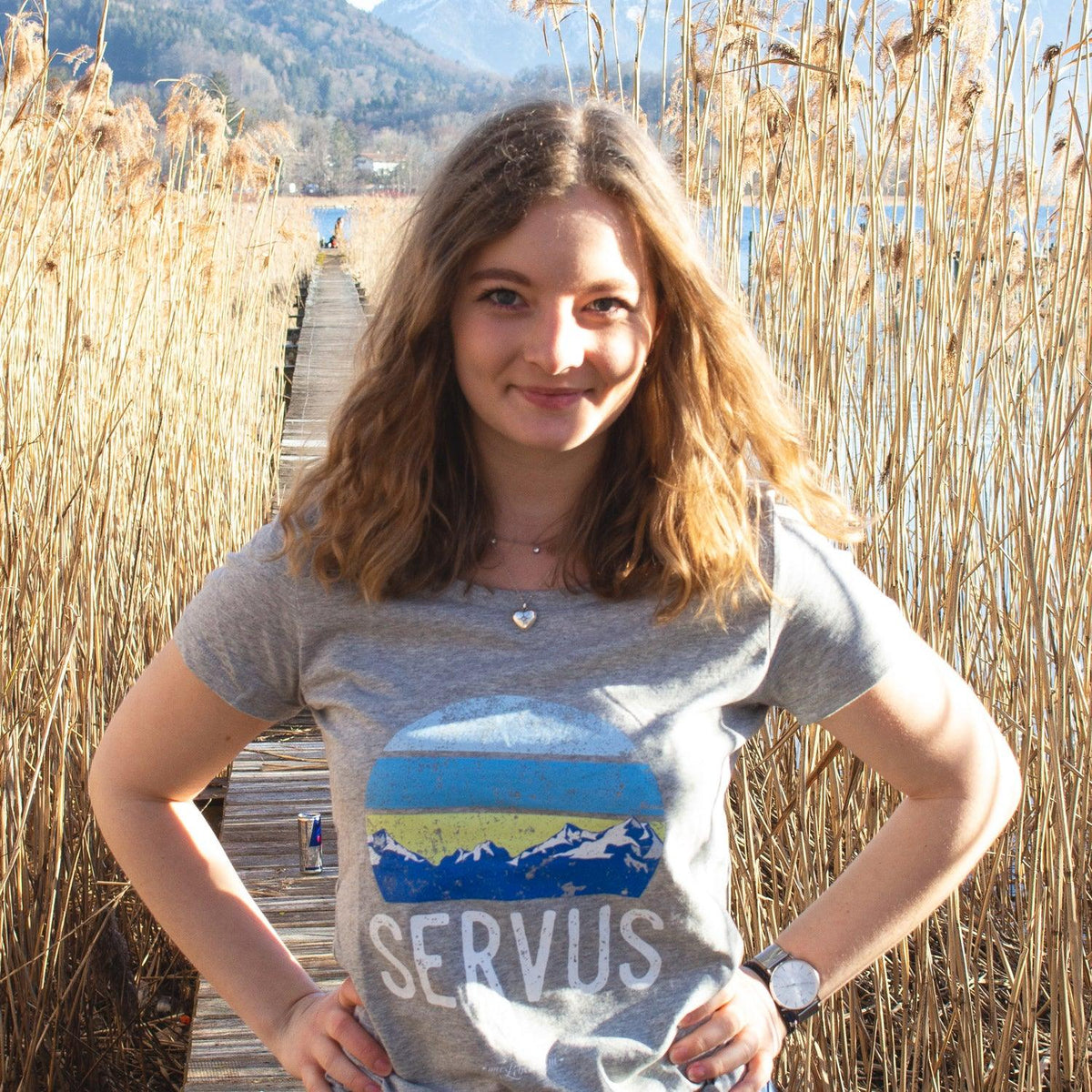 Junge Frau im grauen Damen T-Shirt Servus mit Bergmotiv vor Schilf und See unter blauem Himmel.