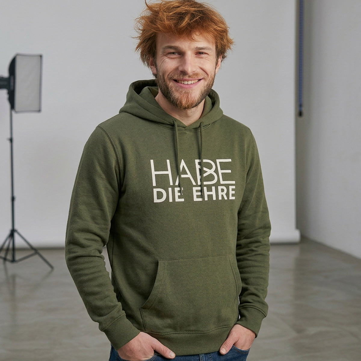 Lächelnder Mann mit rotem Haar trägt olivgrünen Hoodie Habe die Ehre Aufdruck und Kapuze.