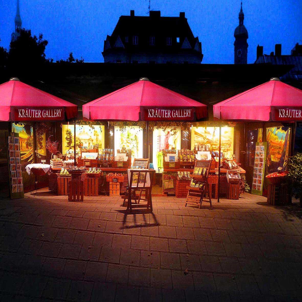 Drei beleuchtete Marktstände mit roten Schirmen vor dunklem Gebäude und Himmel am Abend, bunte Produkte ausgestellt.