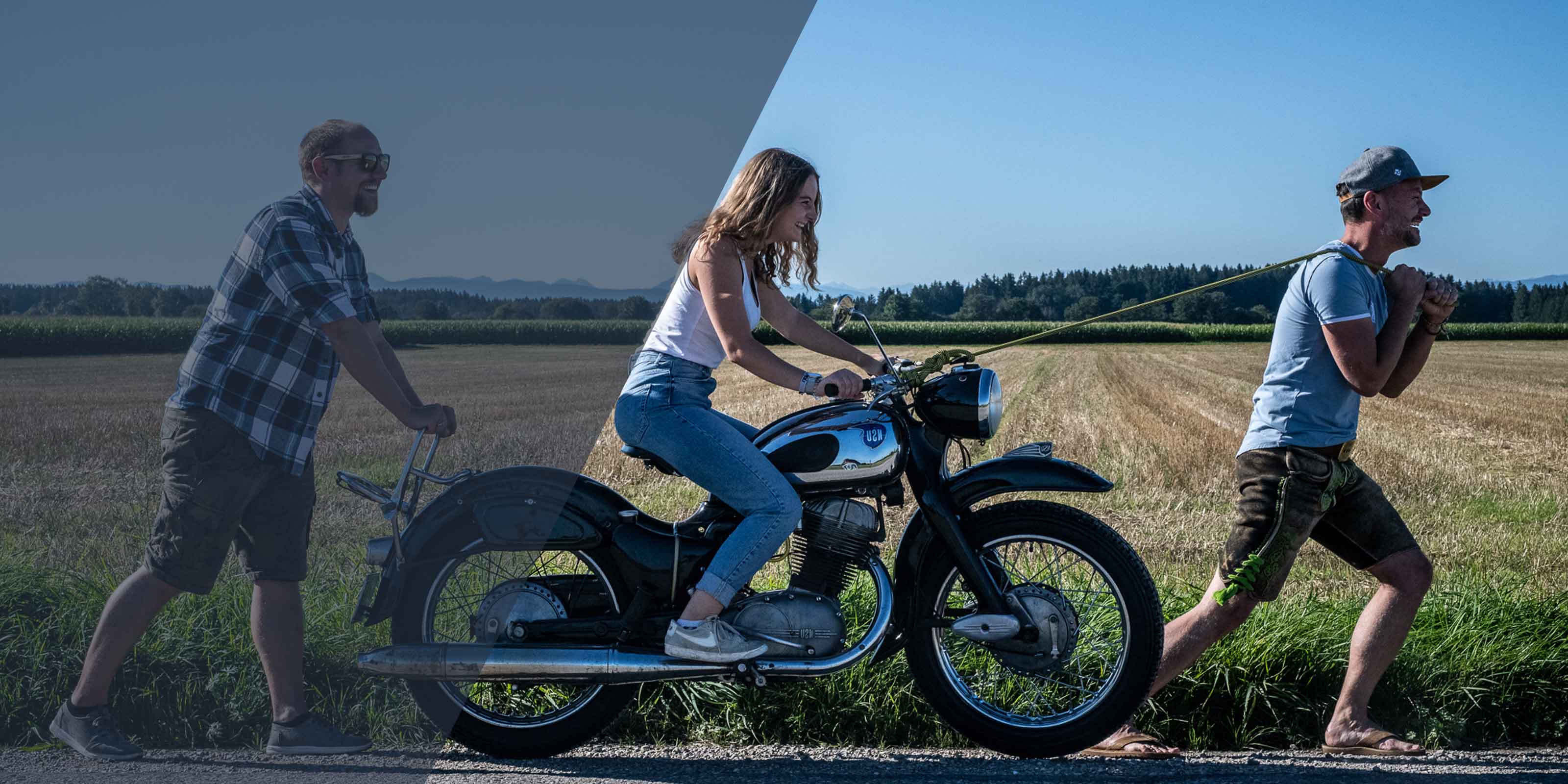 Zwei Männer schieben und ziehen eine Frau auf einem schwarzen Vintage-Motorrad am Rand eines Feldes.