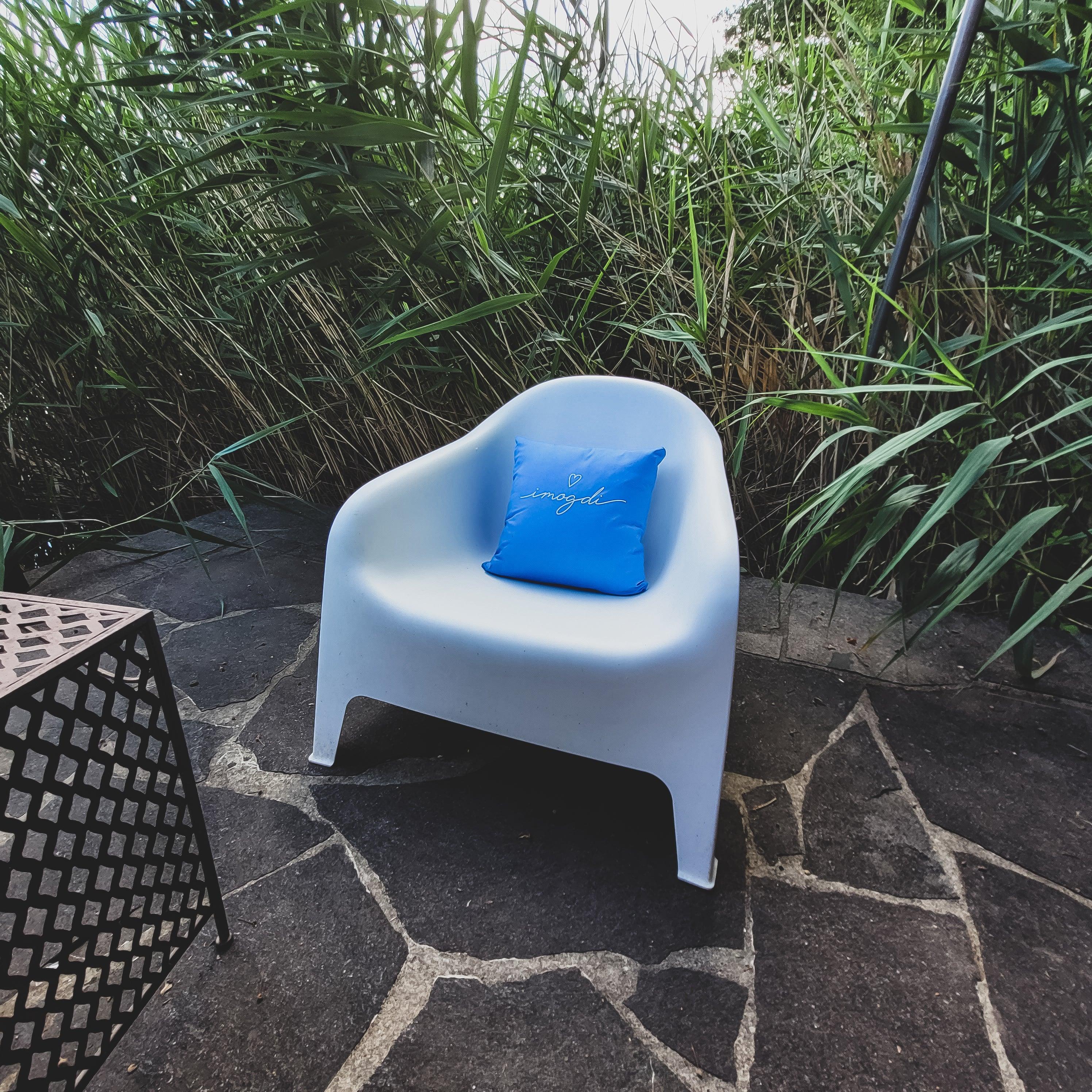 Kissen I mog di in leuchtendem Blau auf weißem Sessel im Garten, umgeben von grünen Pflanzen.