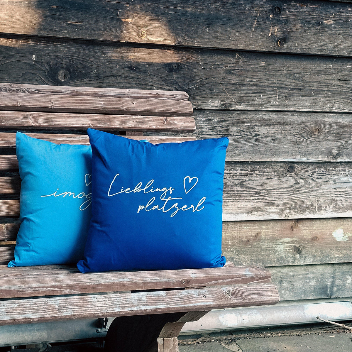 Zwei blaue Kissen "Lieblingsplatzerl" mit weißer Schrift auf einer rustikalen Holzbank vor Holzwand.