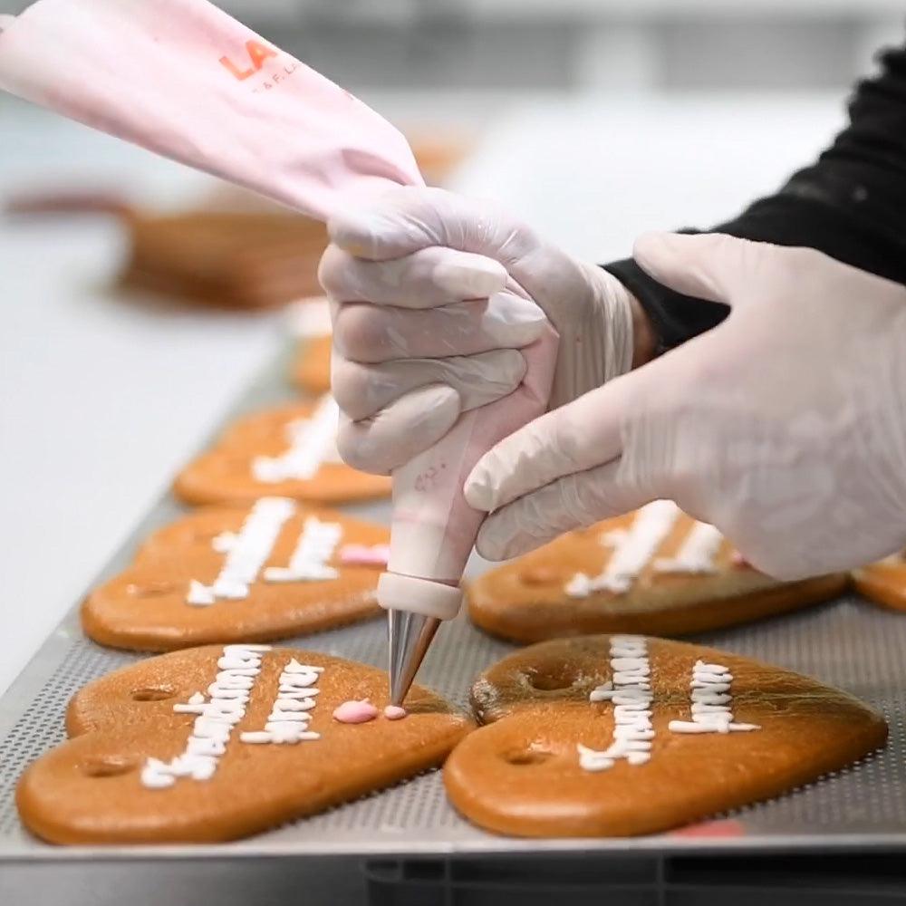 Hände in weißen Handschuhen verzieren Lebkuchenherzen mit rosa und weißem Zuckerguss auf einem Backblech.