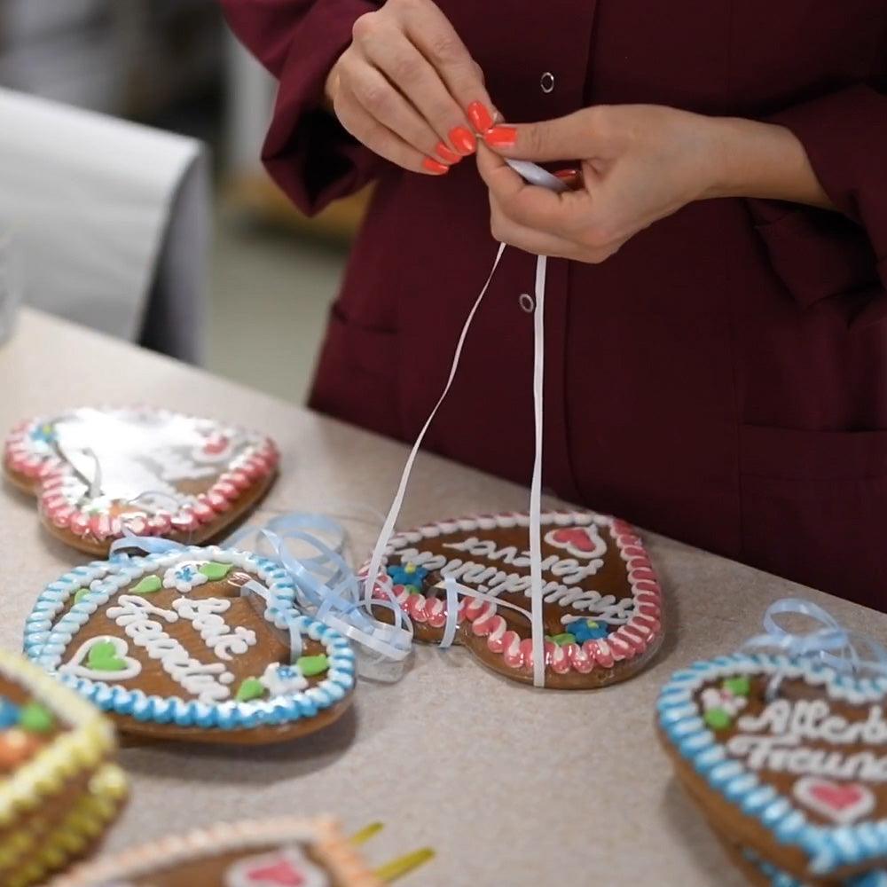 Hände binden ein weißes Band an ein verziertes Lebkuchenherz mit der Aufschrift "Allein für immer".