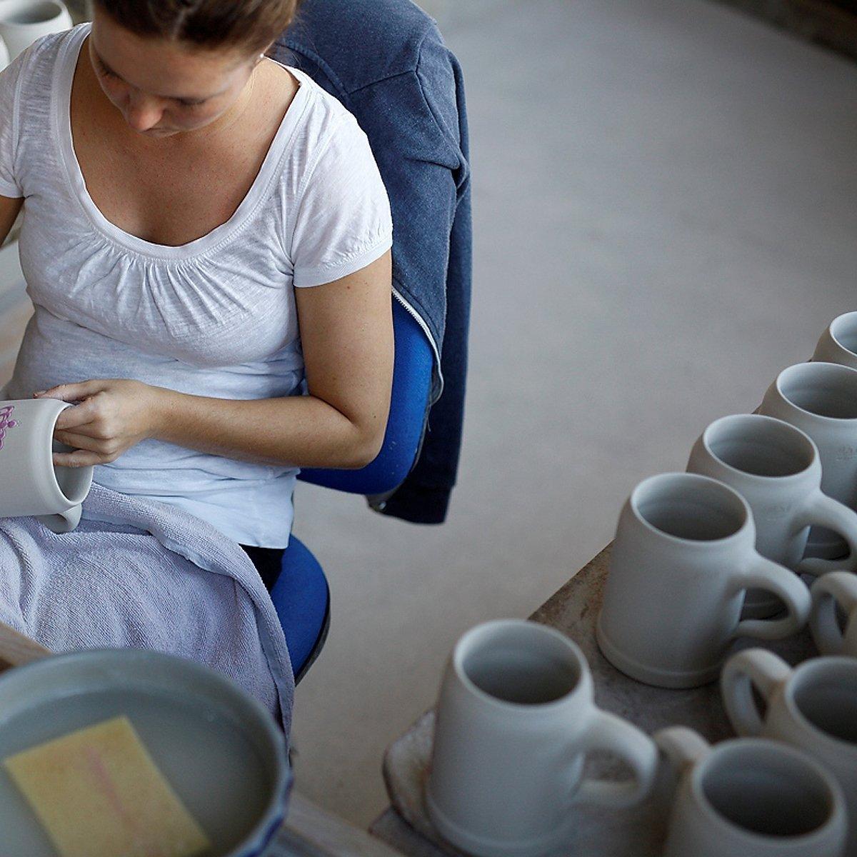 Frau dekoriert einen weißen Keramik Bierkrug von Hand mit lila Details, weitere Krüge warten daneben.