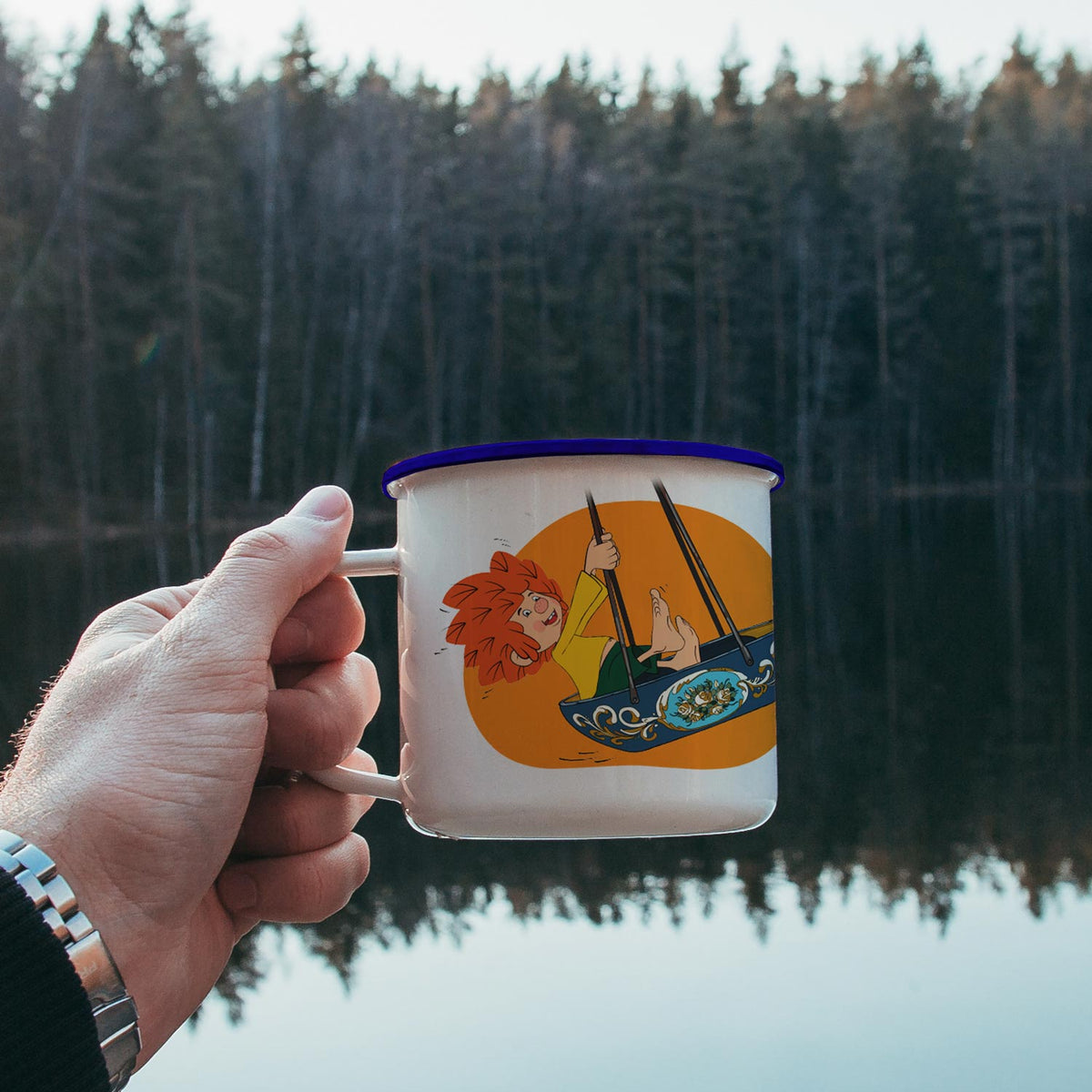 Hand hält weiße ®Pumuckl Emaille-Tasse "Schiffschaukel" mit rotem Pumuckl vor Waldsee.