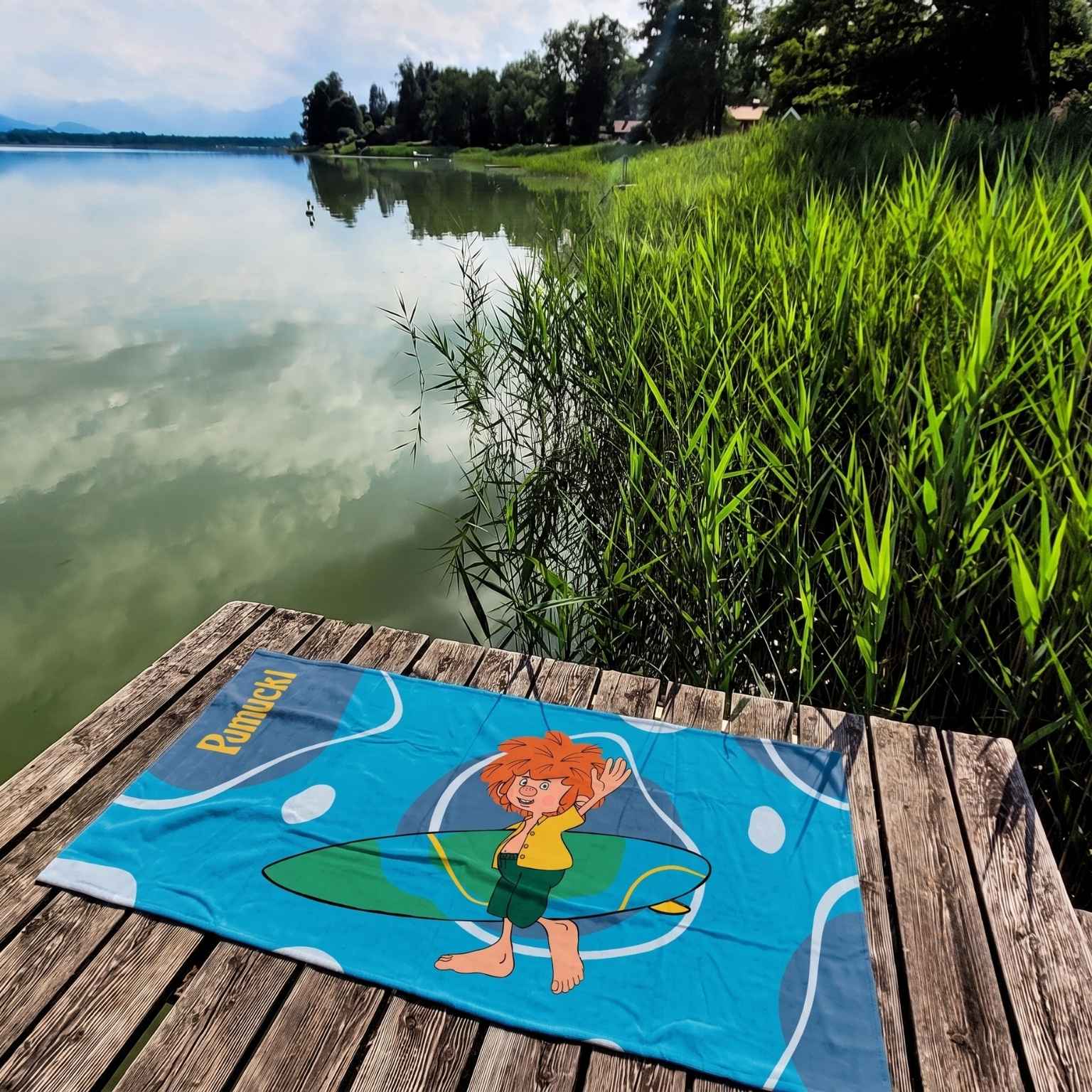 Blaues Pumuckl Handtuch XXL mit Surfbrett-Motiv auf Holzsteg am grünen Seeufer mit Schilf.