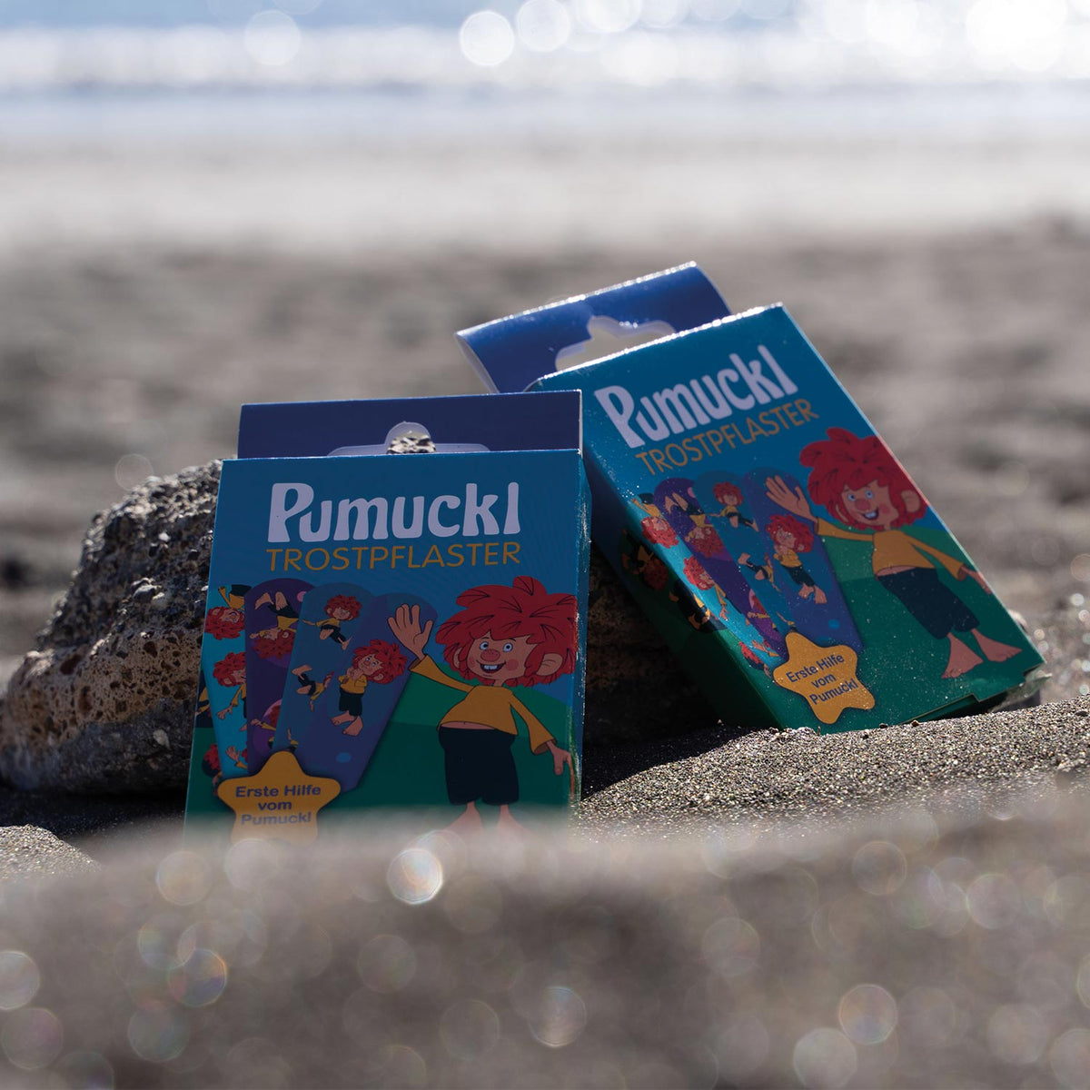 Drei Packungen Pumuckl Trost-Pflaster mit Pumuckl-Design auf dunklem Sandstrand vor glitzerndem Meer.