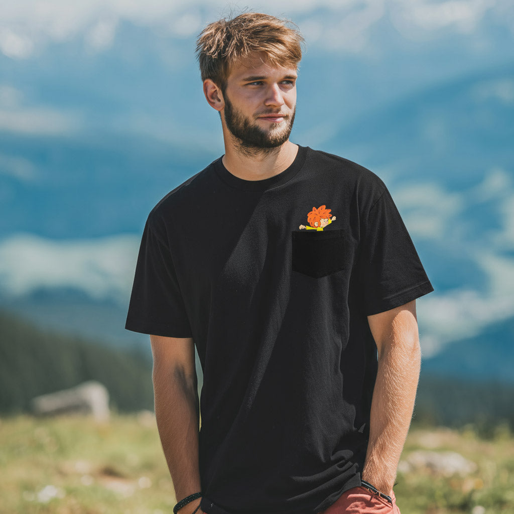 Junger Mann trägt schwarzes ®Pumuckl T-Shirt "Nei da" mit Pumuckl in Brusttasche vor Berglandschaft.