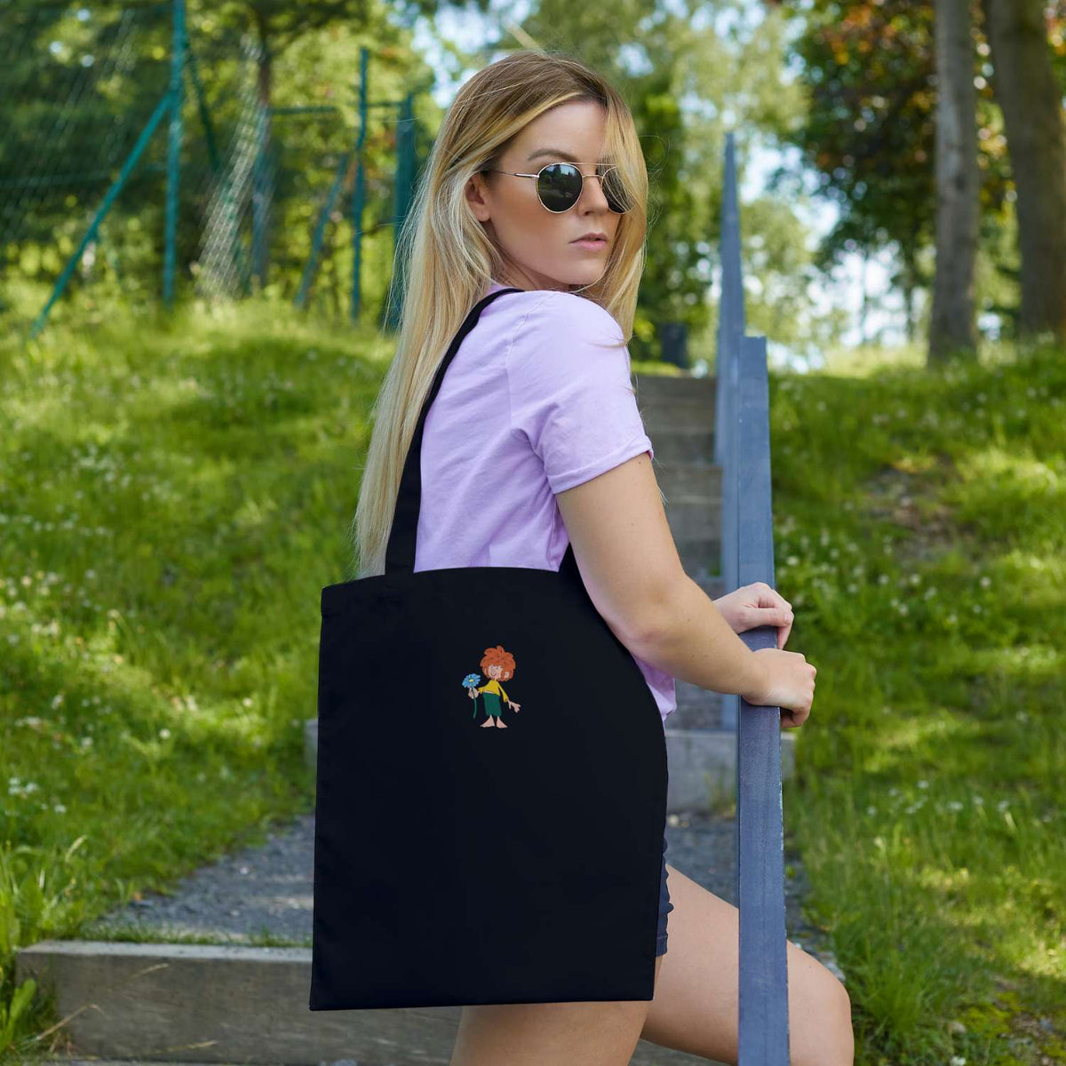Junge Frau mit Sonnenbrille trägt schwarze Pumuckl Tasche Schabernack mit farbigem Kobold-Motiv auf Treppe.