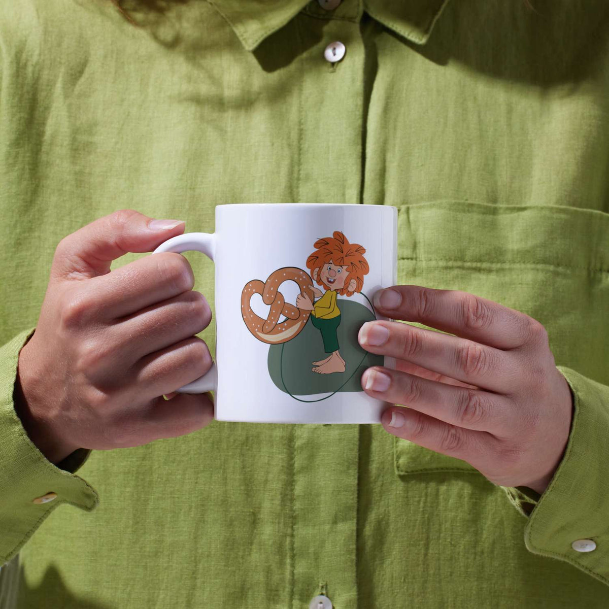 Pumuckl Tasse „Breze“ in zwei Händen vor grünem Hemd
