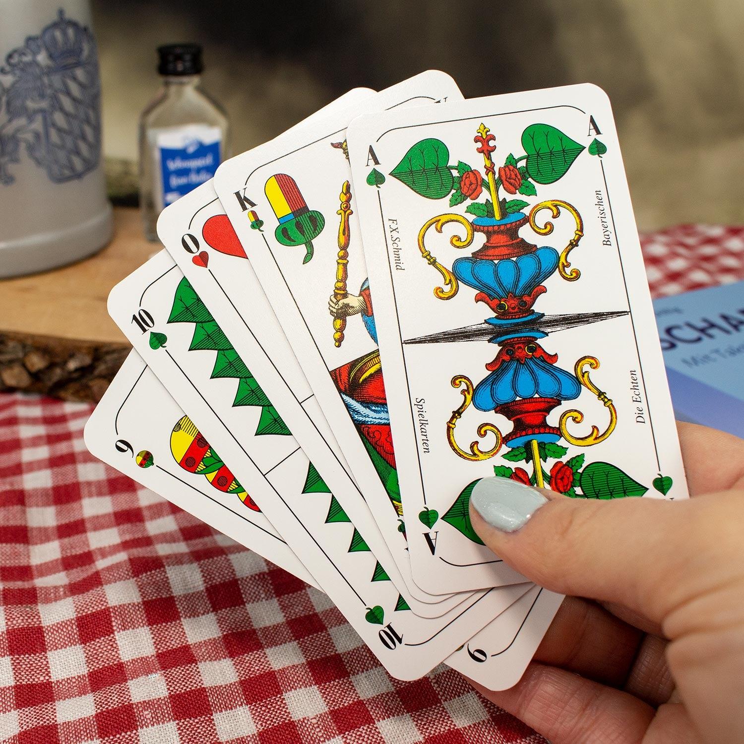 Hand hält bayerische Spielkarten, umgeben von einem roten karierten Tischtuch und traditionellen Accessoires.