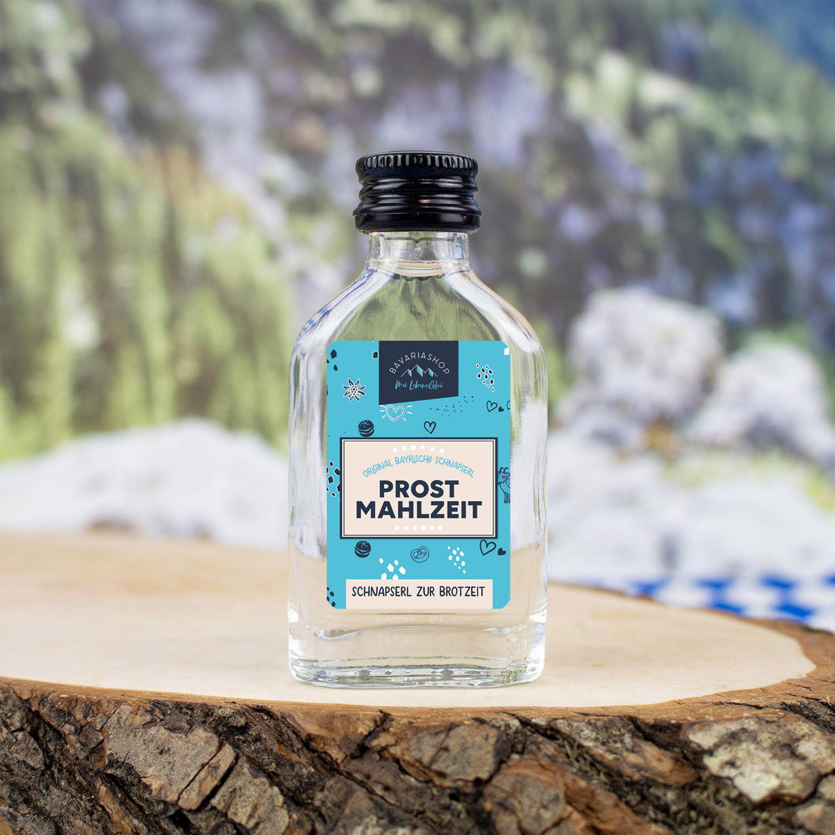 Klare Mini-Schnapsflasche Schnapserl "Prost Mahlzeit!" auf rustikaler Baumscheibe mit Bergkulisse.