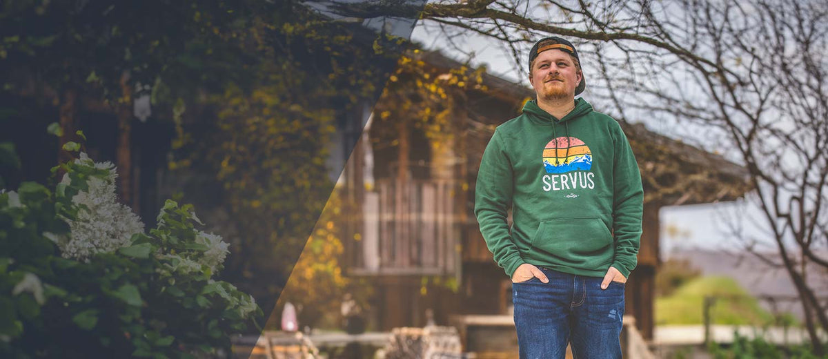 Mann mit grünem bayerischen Hoodie und "Servus" Aufdruck, Jeans und Basecap, im Freien vor rustikalem Haus.