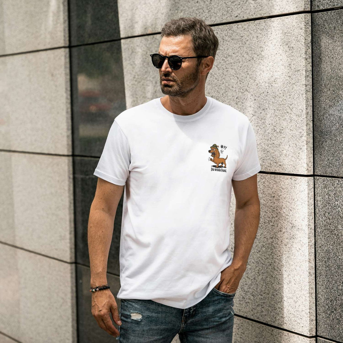 Mann trägt modisches Herren T-Shirt Du wuida Hund in Weiß, mit Dackel-Print und Sonnenbrille vor moderner Wand.