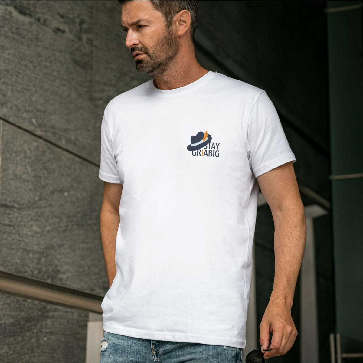 Mann trägt weißes Herren T-Shirt Stay Griabig mit dunkelblauem Hut-Logo und dezenten Nietenjeans.