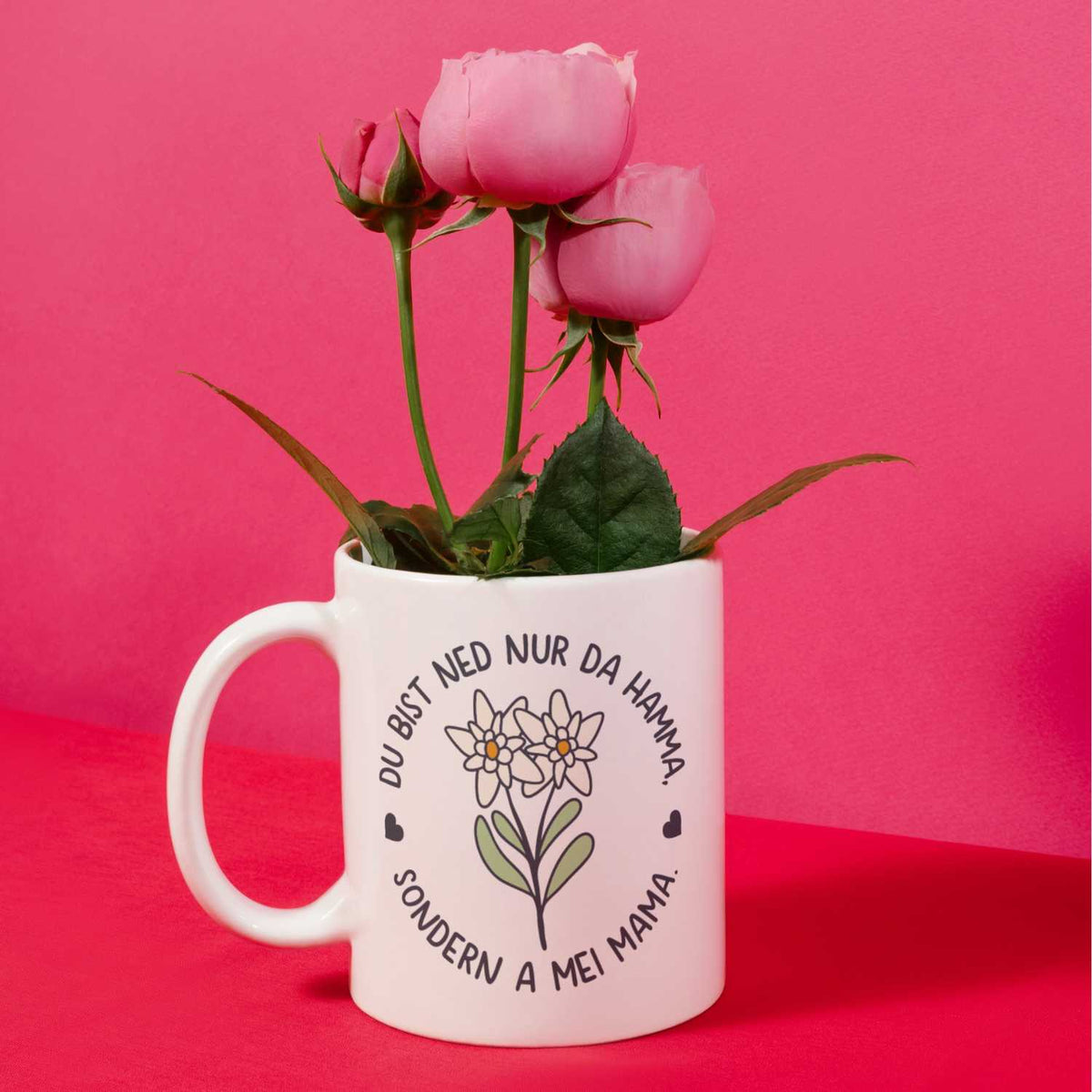 Weißes Kaffee Haferl Hamma Mama mit Wiesenblumen-Motiv und Spruch, gefüllt mit rosa Rosen.