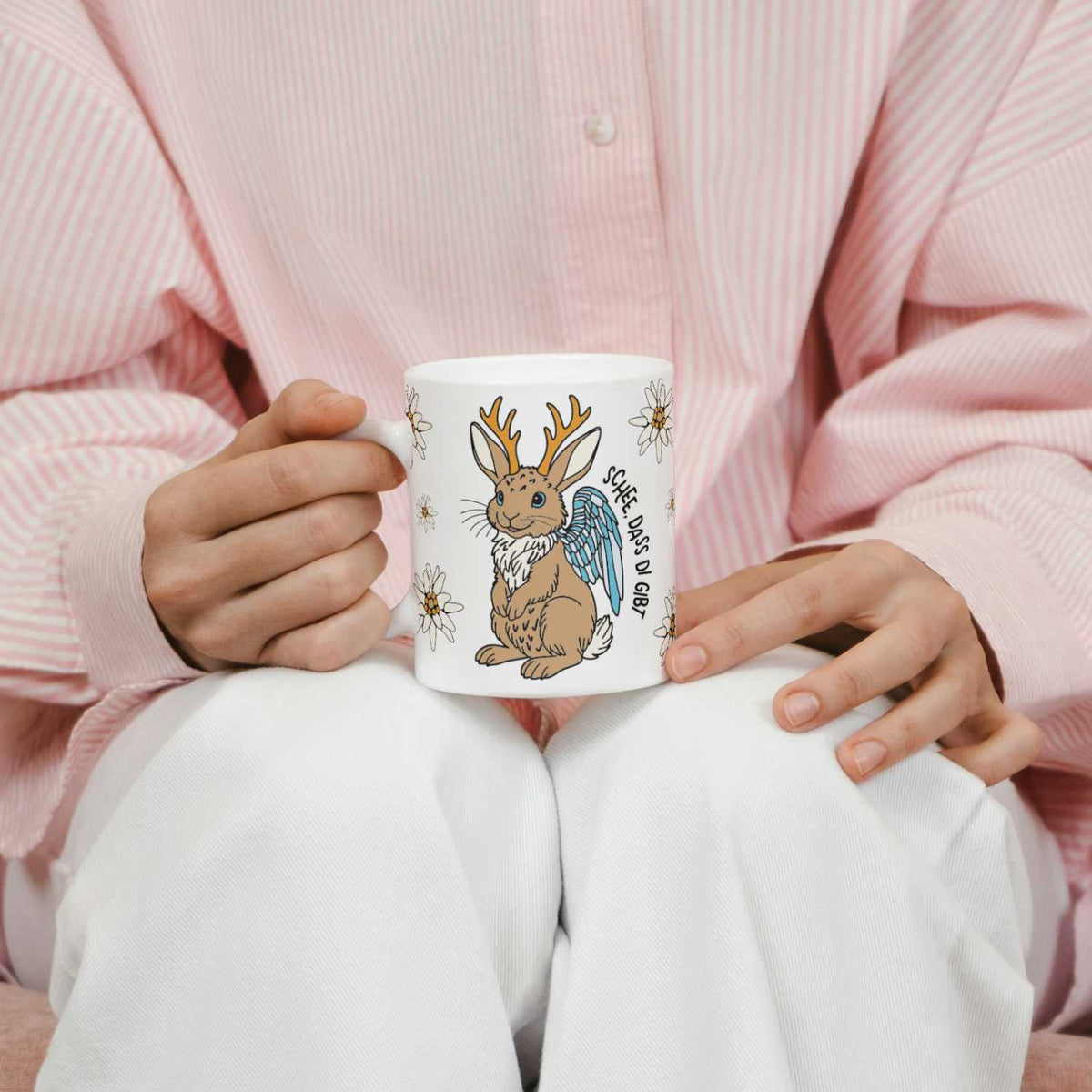 Person hält weißes Kaffee Haferl "Wolpertinger" mit gezeichnetem Hasen mit Geweih und Flügeln.