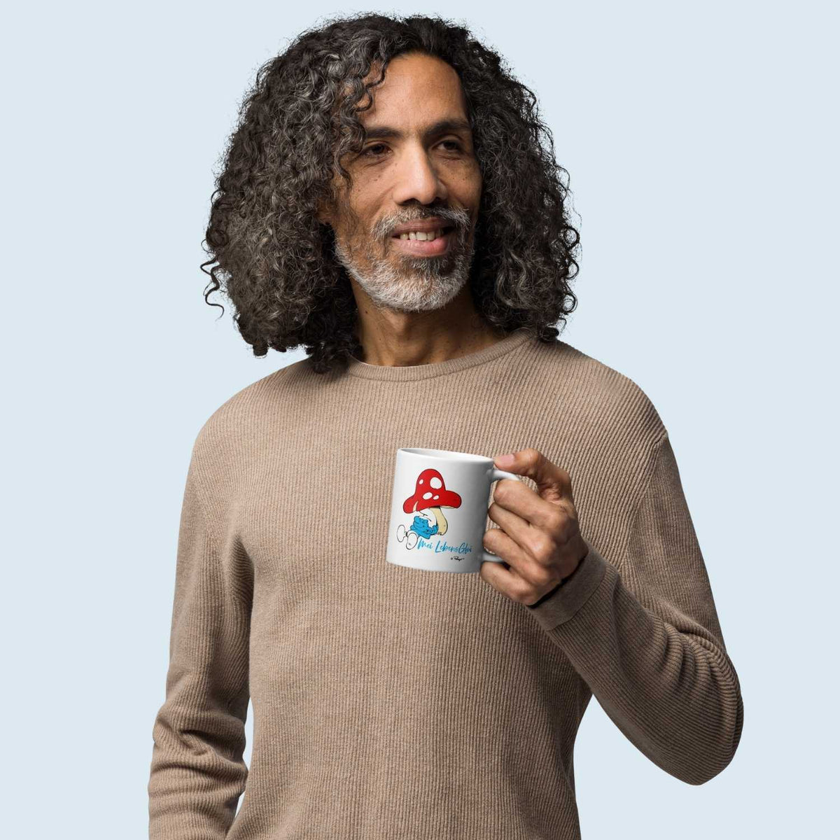 Mann mit krausem Haar hält ®Die Schlümpfe Tasse "Do legst di nieda" mit Schlumpf und Pilzmotiv in der Hand.