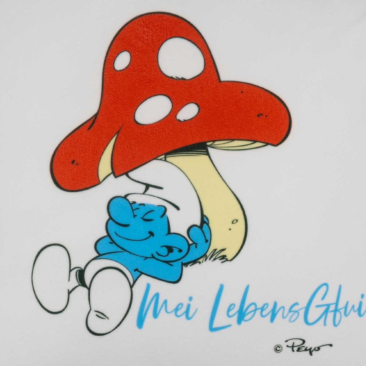Die Schlümpfe "Do legst di nieda!" Kuschelkissen zeigt einen entspannten Schlumpf unter einem roten Pilz.