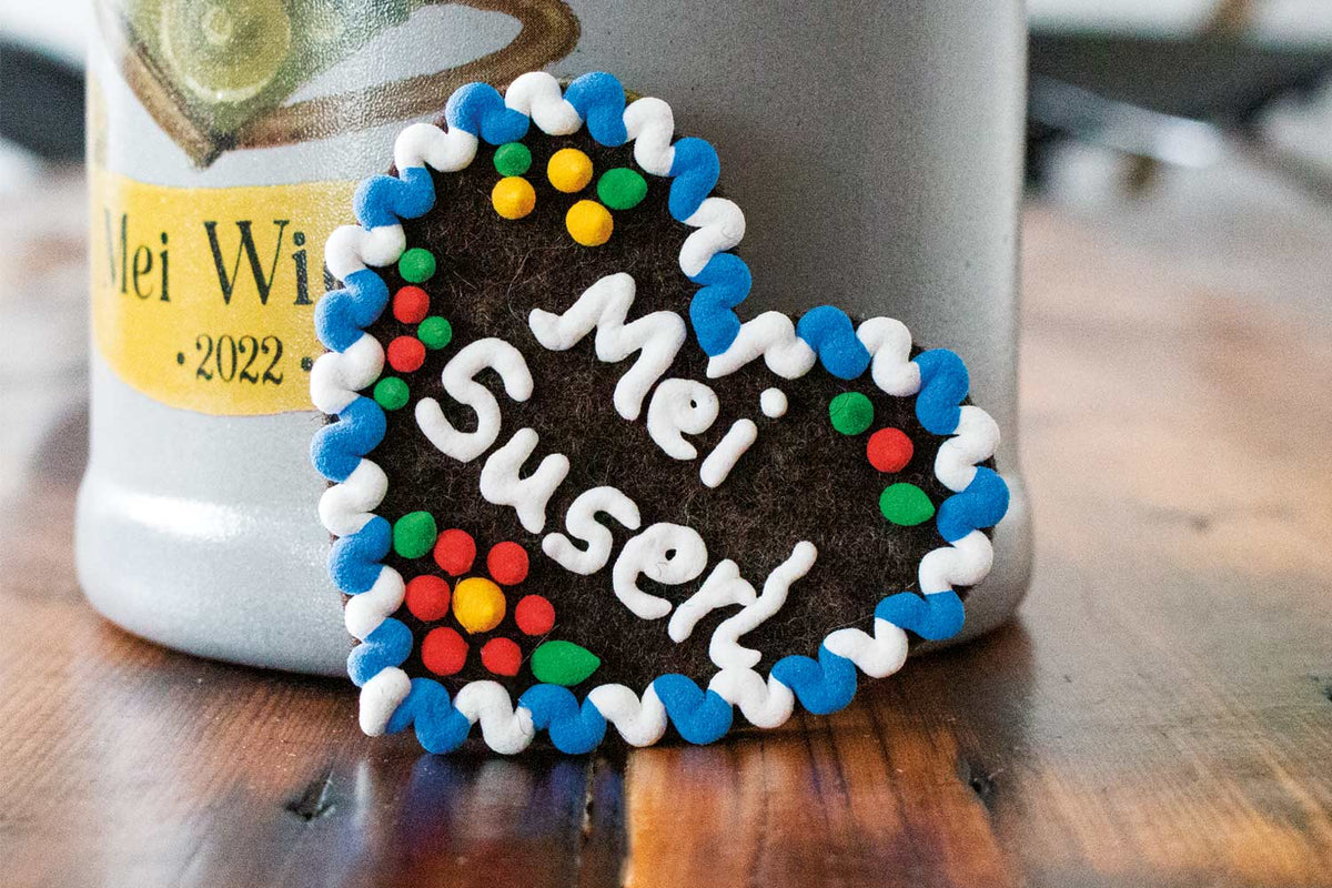 Lebkuchenherz mit blauer, weißer und bunter Zuckerguss-Verzierung und dem Schriftzug "Mei Suserl" neben einem traditionellen Maßkrug.