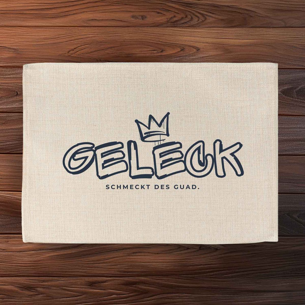 Helles Tischset Geleck mit dunkelblauem Aufdruck Krone und Schriftzug "Geleck schmeckt des guad" auf Holzuntergrund.