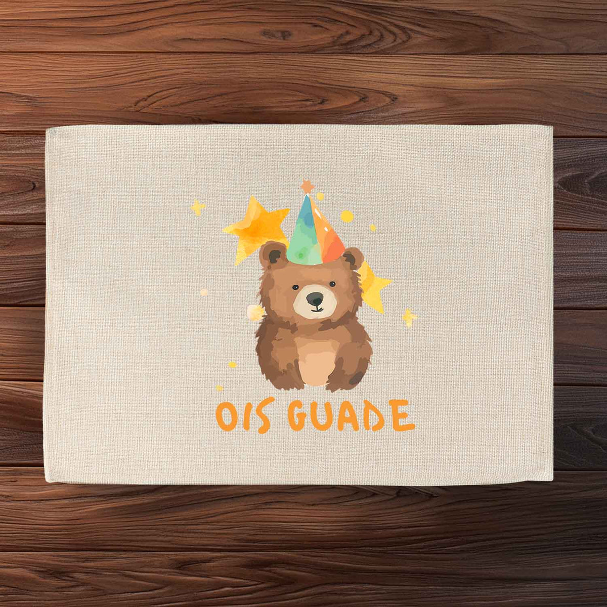 Beiges Tischset "Ois Guade Bär" mit niedlichem Bären, Partyhut, Sternen und Schriftzug auf Holzuntergrund.