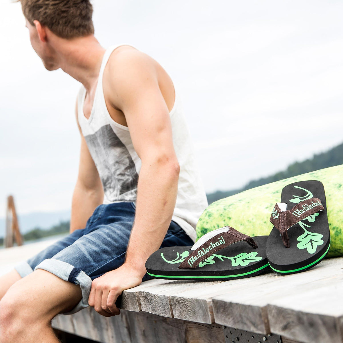 Mann in Tanktop und Jeansshorts auf einem Seesteg neben einem Paar Trachten Flip Flops Haferlschuh mit Eichenlaub-Motiv