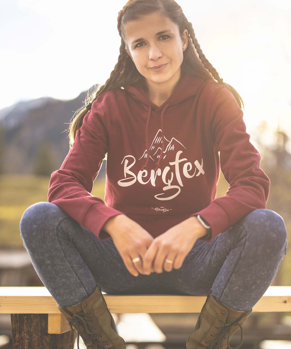 Junge Frau mit geflochtenen Zöpfen in einem bordeauxroten Hoodie mit Bergblick-Logo, sitzend auf einer Holzbank.