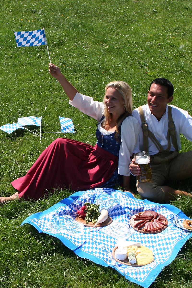Lachendes Paar in Tracht beim Picknick auf bayerischer Decke, mit Bierkrug, Wurstaufschnitt und Käse auf Holzbrettern.