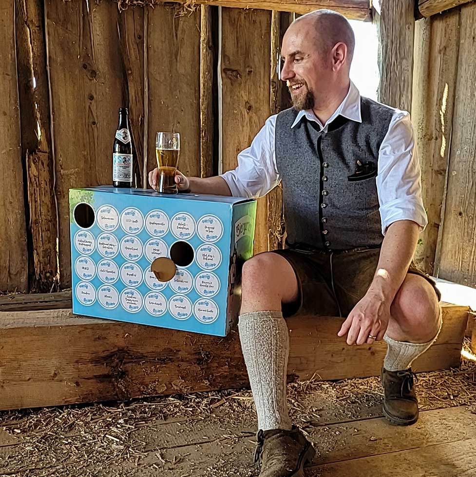 Traditionell gekleideter Mann mit Lederhose und Trachtenweste, sitzt lächelnd mit einem Bierglas neben einer blauen Geschenkbox.