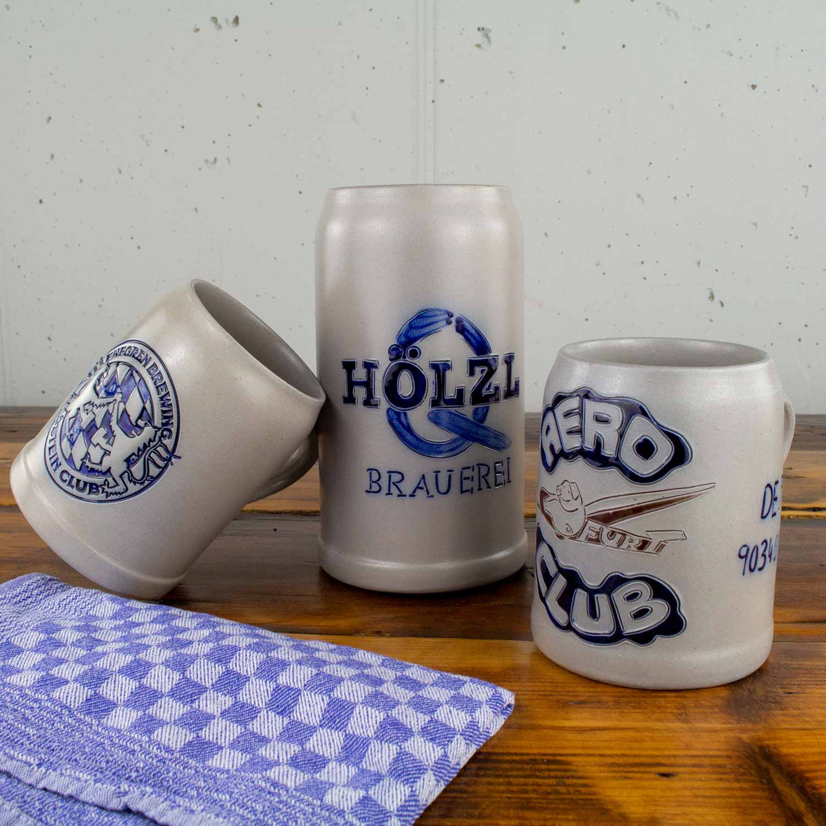 Drei traditionelle Bierkrüge aus Steingut mit detaillierten blauen Emblemen, darunter Brauerei- und Club-Logos, auf einer Holzfläche mit kariertem Tuch.