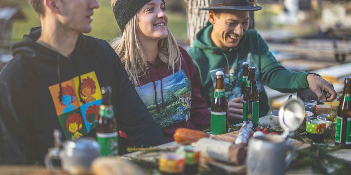 Drei junge, lachende Freunde am Biertisch im Grünen, mit Hoodies und bayerischen Bierkrügen.