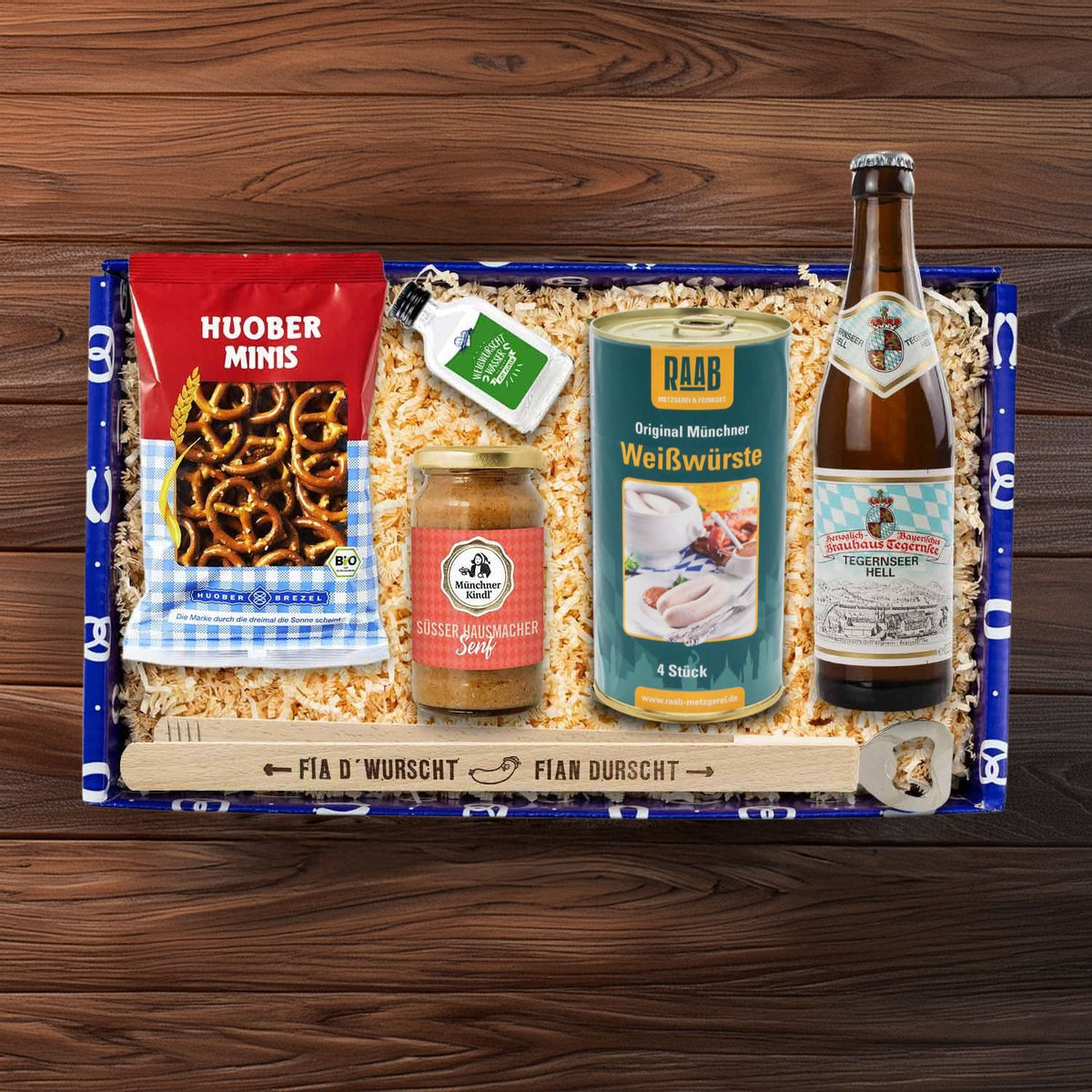 Geschenkbox mit Münchner Weißwürsten, süßem Senf, Brezeln, Bier und "Weißwurschtwasser" auf Holztisch.