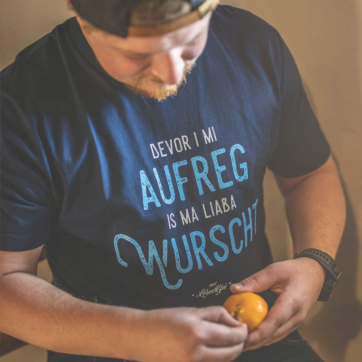 Herren T-Shirt Bevor i mi aufreg... in Dunkelblau mit hellem Text, getragen von einem Mann mit Bart, der eine Mandarine hält.