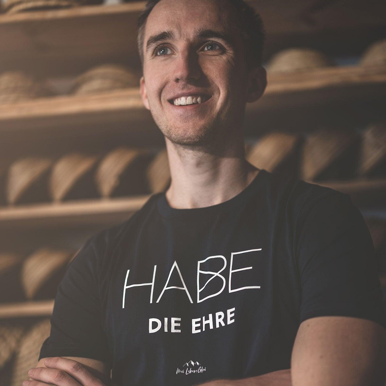 Lachender Mann im schwarzen Herren T-Shirt Habe die Ehre in natürlicher Umgebung mit Holzregal Hintergrund.