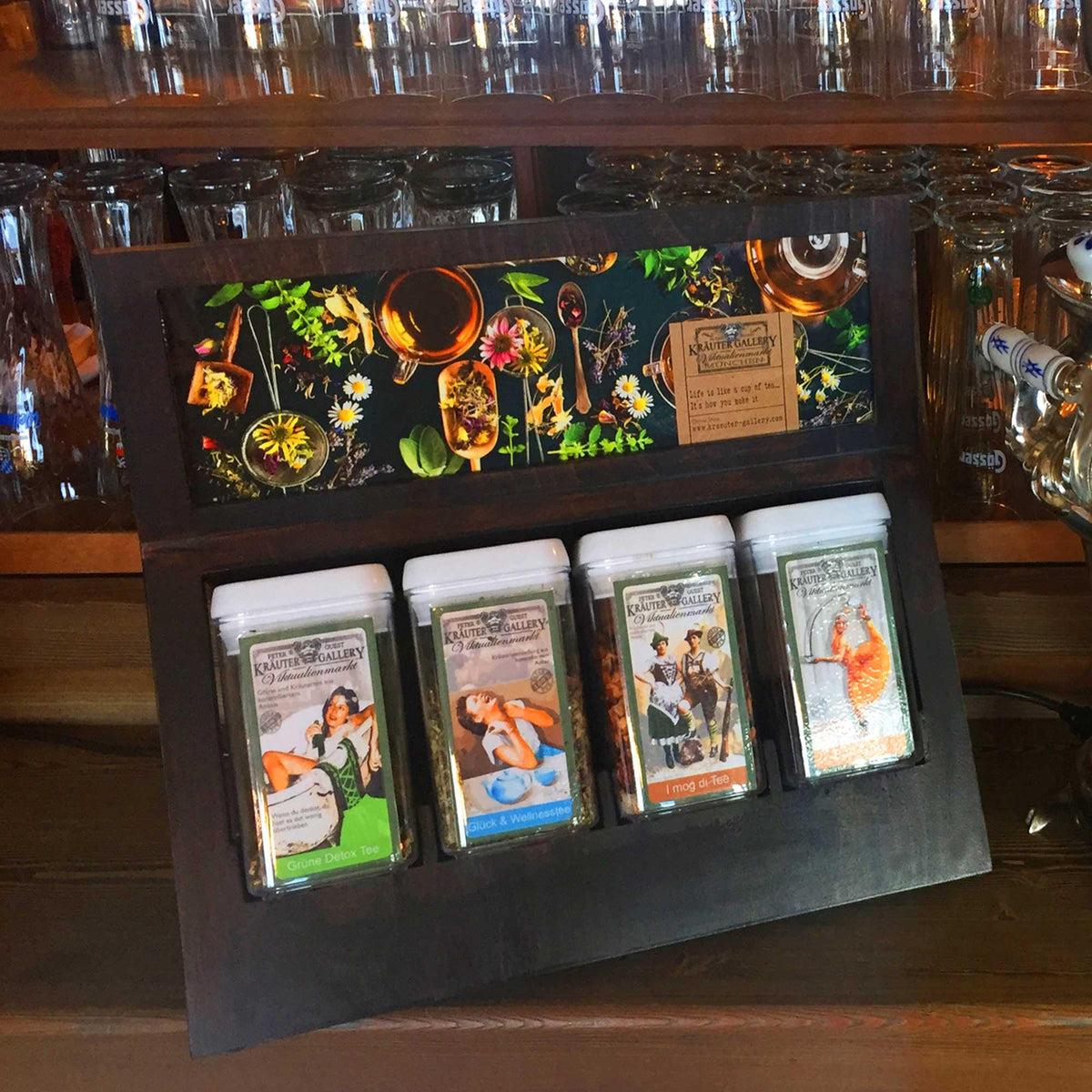 Holzdisplay mit vier verschiedenen Kräutertee-Sorten in transparenten Dosen, darüber Kräuter-Dekor.