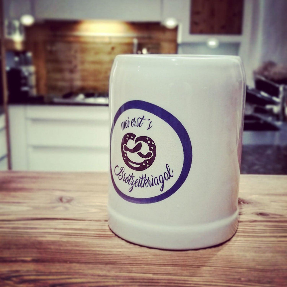 Weißer Keramikkrug mit blauem "mei erst's Brotzeitkriagal" Logo und Brezel-Motiv auf rustikaler Holztheke.